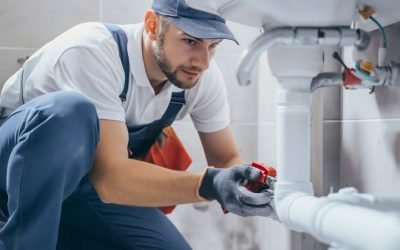 Young handsome plumber wearing a working uniform and a cap, kneeling down in the bathroom to fix or repair the white pipes under the sink. Home service or maintenance, professional handyman job